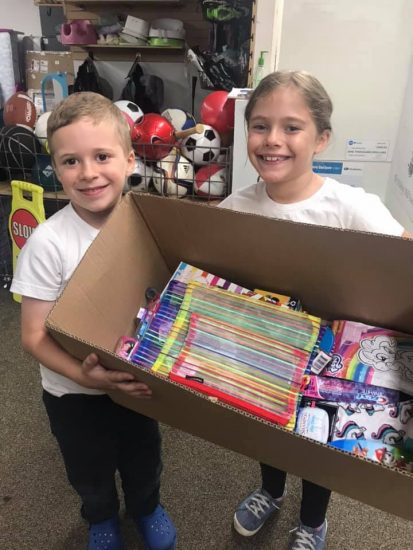 A smiling young boy and girl hold a large box between them, full of items donated for foster kids.