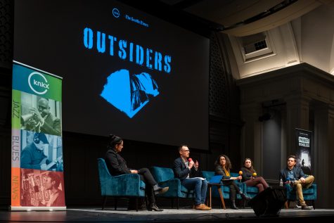 Five people sit in chairs on a stage under a screen showing the "Outsiders" podcast logo.