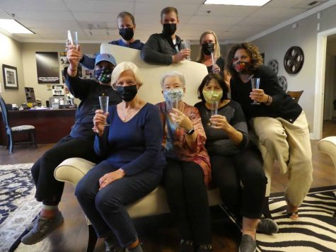 Eight people, wearing COVID masks, sit on a couch, raising champagne glasses.